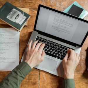 High-angle view of hands typing on a laptop surrounded by books and papers.